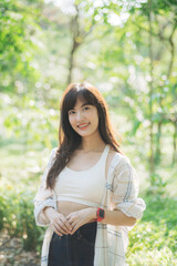 cheerful young woman in casual attire stands outdoors in lush green park, smiling warmly under soft sunlight