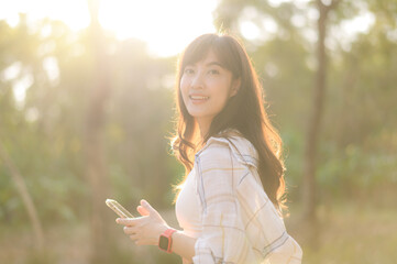 Naklejka premium Asian woman enjoying summer sunlight while breathing fresh air in serene outdoor setting, holding smartphone with cheerful expression