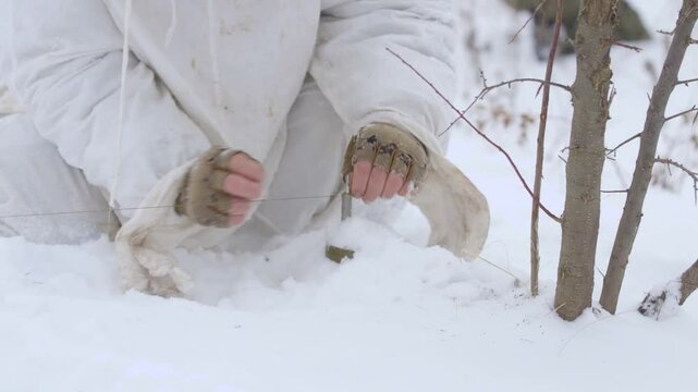 Sapper soldier places an anti-personnel mine explosive on a tripwire. Military sapper mines and camouflages a mine explosive in snow. Unrecognizable soldier in a winter camouflage suit mines an area.