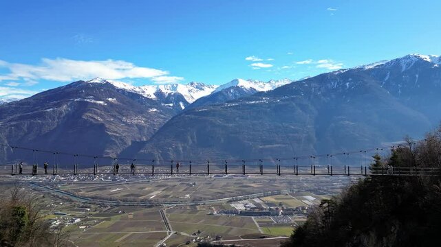 Saillon, Switzerland - 15 February 2025: Tourists Walking Across the Passerelle &agrave; Farinet, a Wooden Bridge Above the Salentse Gorge with Views of the Swiss Alps on a Beautiful Sunny Day