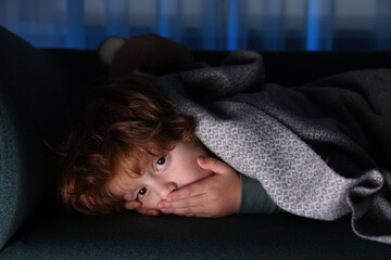 Afraid boy hiding under blanket on sofa at night
