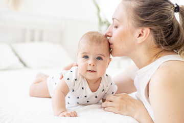 a young mother gently kisses and hugs her newborn baby lying on a light white bed at home, the concept of a young family and motherhood