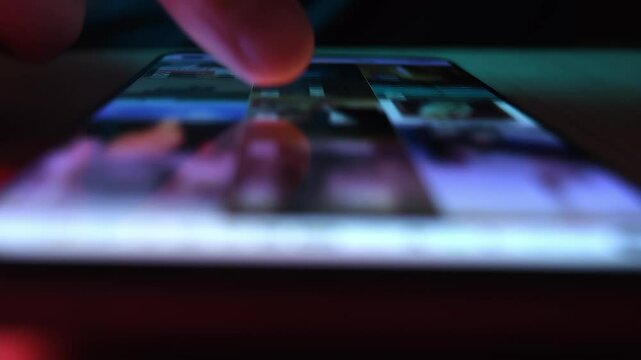 Finger moves across phone screen in dim setting; it unveils a vivid social feed and highlights late-night digital immersion