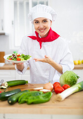 Smiling female chef in white uniform offering plate of vegetable salad in home kitche