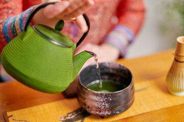 Green teapot pouring steaming water into black ceramic bowl during traditional tea preparation, emphasizing simplicity and cultural mindfulness