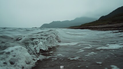 A gently rolling wave greets the foggy shore, encapsulating the serene yet powerful presence of the sea against muted, cloudy cliffs.