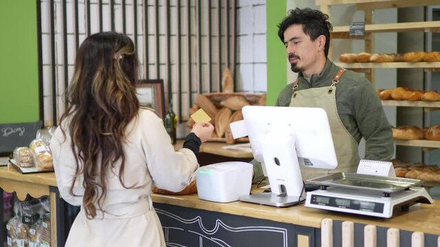 Baker processing credit card payment from customer at local artisan bakery, exchanging transaction at point of sale while displaying fresh baked goods
