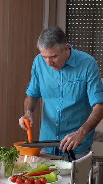 Casual Middle-aged Man with Gray Hair, Slices Carrots at Home on a Table full of Various Vegetables. The video is perfect for promoting healthy food, food tutorials and lifestyle of middle-aged men.