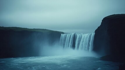Fototapeta premium A powerful waterfall cascades over a cliff, enveloped in mist and framed by lush greenery under an overcast sky.