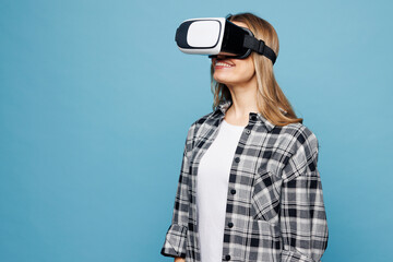 Side profile view young smiling woman wears checkered grey shirt casual clothes watching in vr headset pc gadget isolated on plain pastel light blue cyan background studio portrait. Lifestyle concept.