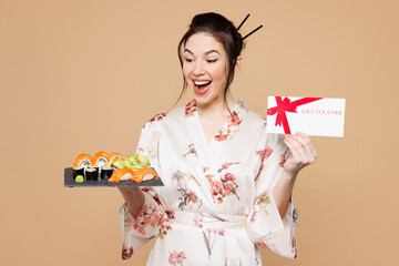 Young surprised shocked woman wearing kimono clothes hold eat raw fresh sushi roll served on black plate gift certificate coupon voucher card isolated on plain beige background. Japanese food concept.