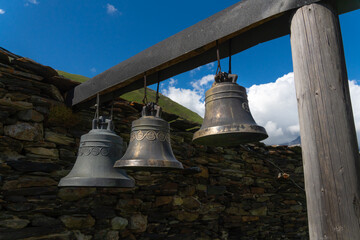 Three large bells hang from a wooden beam in front of a stone wall, with green mountains and a blue sky in the background, creating a serene and traditional scene