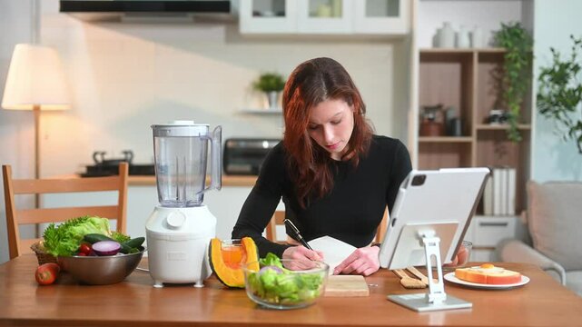 Young female in kitchen and writing down healthy recipe or daily ration diet at home. Active healthy lifestyle
