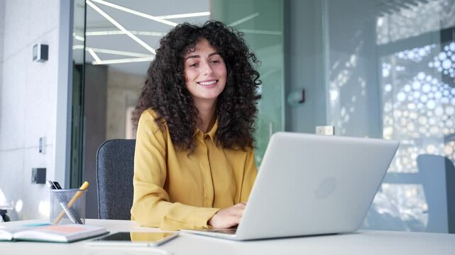 Young adult female employee typing on laptop while sitting at workplace in business office. A smiling curly brunette woman works on a computer, chats online or communicates remotely with a client