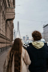 A young couple walks arm in arm through a city street on a cold day, surrounded by historic architecture and urban scenery.