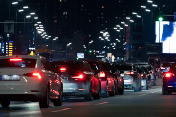 A lively and bustling city street at nighttime, featuring a significant traffic jam with the brilliant glow of numerous tail lights alongside vibrant billboards illuminating the background