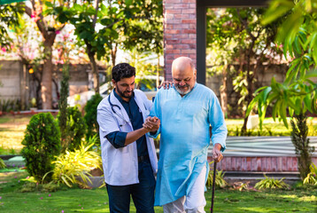Elderly Indian patient using a walking stick walks with a doctor in a hospital park, discussing...