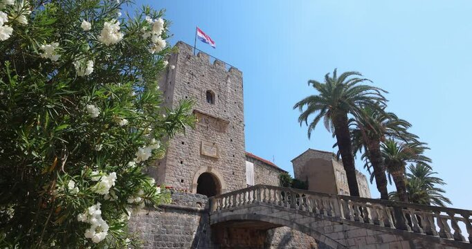 Korcula town in island of Korcula in Adriatic sea in Croatia, Europe. Historic stone tower, crowned with a flag, rises amidst lush greenery and palm trees against a clear blue sky.