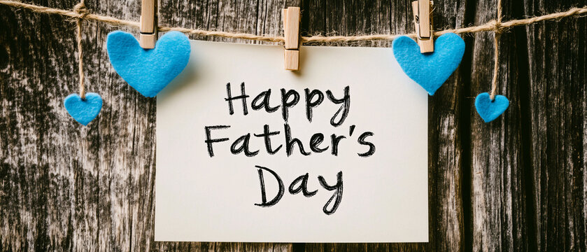 A rustic Father&rsquo;s Day greeting card with handwritten text hangs on a wooden background decorated with small blue hearts on a string  
