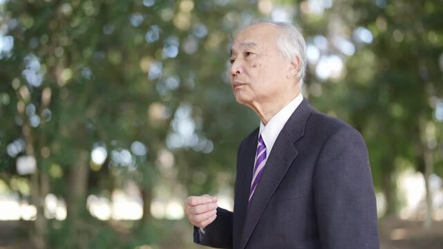 A 78-year-old Japanese man in a suit stands in a natural park filled with trees and talks.