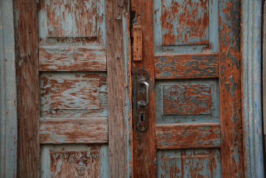 Photo of old double wooden doors with cracked, peeling paint in faded turquoise blue and rusty orange-brown tones. Carved recessed panels, aged wood grain and a vintage metal handle with a lock plate 