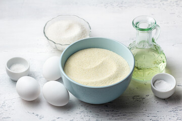 Baking ingredients: light blue bowl with semolina, fresh white eggs, a glass bowl of granulated sugar, and a bottle of vegetable oil on a white kitchen surface