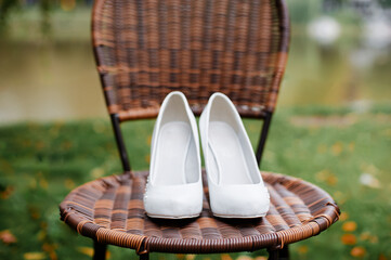 A pair of elegant white high heels shoes displayed on a wicker chair in a serene outdoor setting....