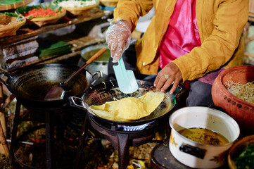 A person cooking traditional street food in a wok at a market, using a spatula to flip a crepe-like dish. Various ingredients and cooking utensils are visible around the cooking area.