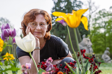 Smiling woman with red hair enjoying colorful flowers in a garden. Red haired mature woman enjoying...