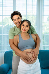 Happy Asian couple man and woman embracing pregnancy with hands forming heart shape on belly, looking at camera