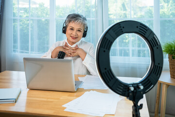 Senior influencer Asian woman using ring light and microphone for online podcast and digital content creation, looking at camera