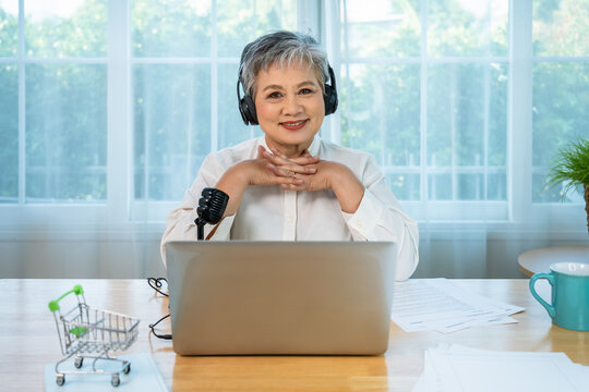 Senior influencer Asian woman hosting an online webinar with a microphone and laptop in a professional home office setup, looking at camera