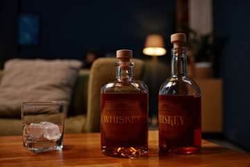 Whiskey bottles arranged on wooden table beside glass with ice cubes in cozy room with dim lighting...