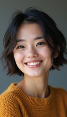 A cheerful young Asian woman with short, wavy hair beams with a bright smile, wearing a cozy mustard sweater. Her joyful expression radiates positivity and warmth, inviting connection and happiness.