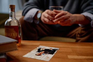 Person sitting on couch holding glass of whiskey, with bottle and photo of a soldier on wooden table, creating emotional and nostalgic atmosphere