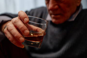 Senior man holding a glass of whiskey with a thoughtful expression. Close-up of hand prominently featuring the glass and capturing the depth of the moment
