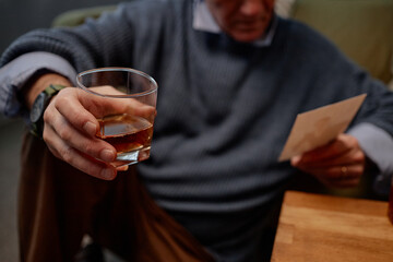 Person seated indoors, holding a glass of whiskey while attentively reading a letter. Image highlights thoughtful and reflective moment captured mid-action
