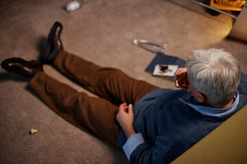 Senior man with grey hair sitting on carpet, holding drink in one hand and book in the other...