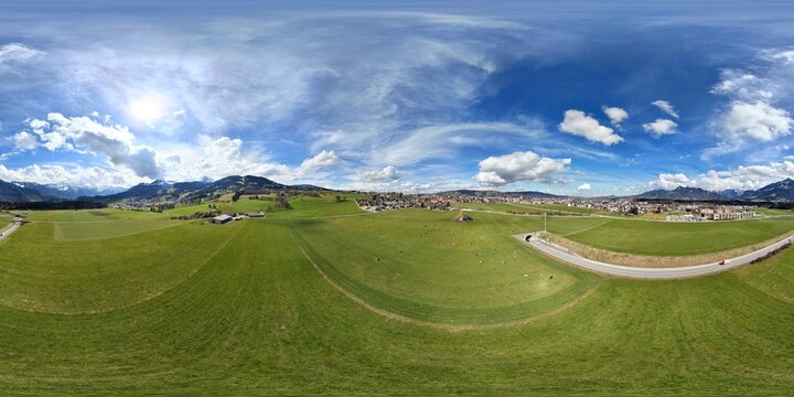 Panoramic aerial view of a rural landscape with green fields, a small town, mountains, and a blue sky with clouds. A road with a tunnel curves through the scene, with cows grazing in open pastures.
