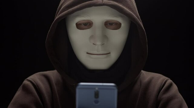 Masked criminal uses phone to commit fraud. Concept of hacking and cybersecurity via smartphone.