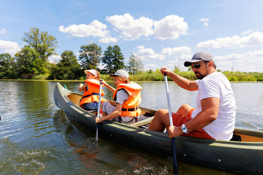 Man with two children enjoy having fun paddling canoe rent across lake river water wearing bright orange life vests hot sunny summer day. Healthy recreational activity lifestyle vacation trip journey