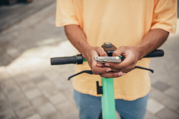 Young African American man using smartphone while riding electric scooter in urban street during sunny day, dressed in casual t-shirt.