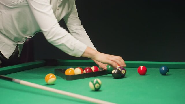 Person in white shirt using triangle rack to arrange billiard balls on green pool table. Cue stick lying nearby, balls scattered before being set for break shot. Dark background