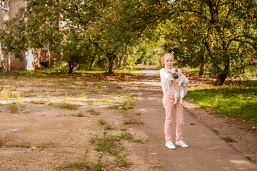 Fototapeta premium Blonde girl in a white poncho holding a small dog in a warm, sunny, old park