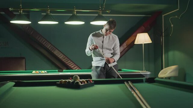 Sportsman in white shirt and grey trousers sits on green pool table, holding cue stick with focused expression. Another pool table with arranged billiard balls is visible in background