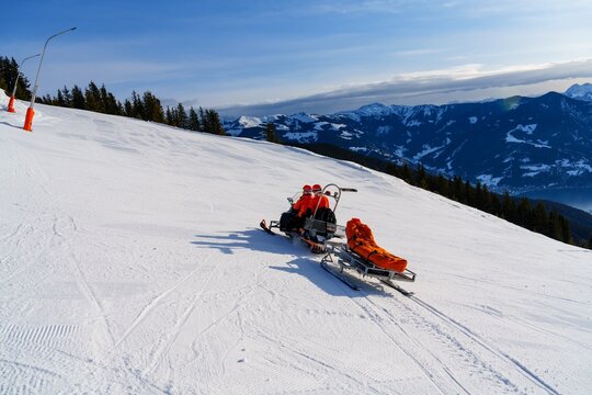A snowmobile rescuer is driving a rescue sled with an injured skier on it. The background is a beautiful mountain landscape with snow-covered trees and mountains.