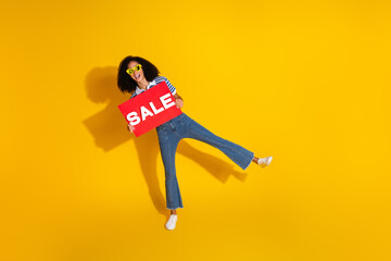 Young stylish woman in casual outfit holding a sale sign against a bold yellow background, expressing excitement and promoting