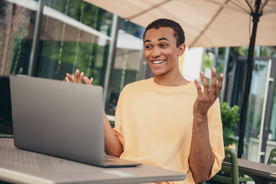 Young man in trendy casual outfit enjoying leisurely work on laptop outdoors, sitting under umbrella on sunny day
