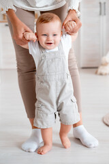 mom teaches the baby to walk at home, the baby boy learns to walk by taking the first steps holding his mother's hand