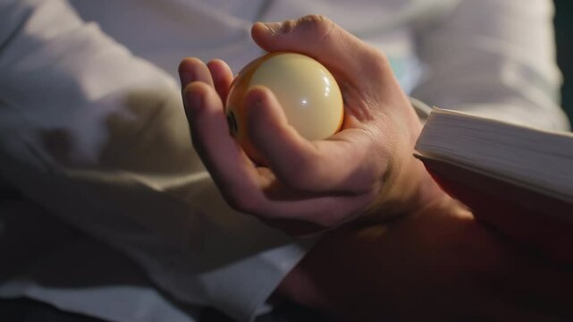 Close-up shot of hand in white shirt, face not visible, holding an open book while rolling a billiard ball in one hand. Focused gesture, moody lighting, and intense concentration